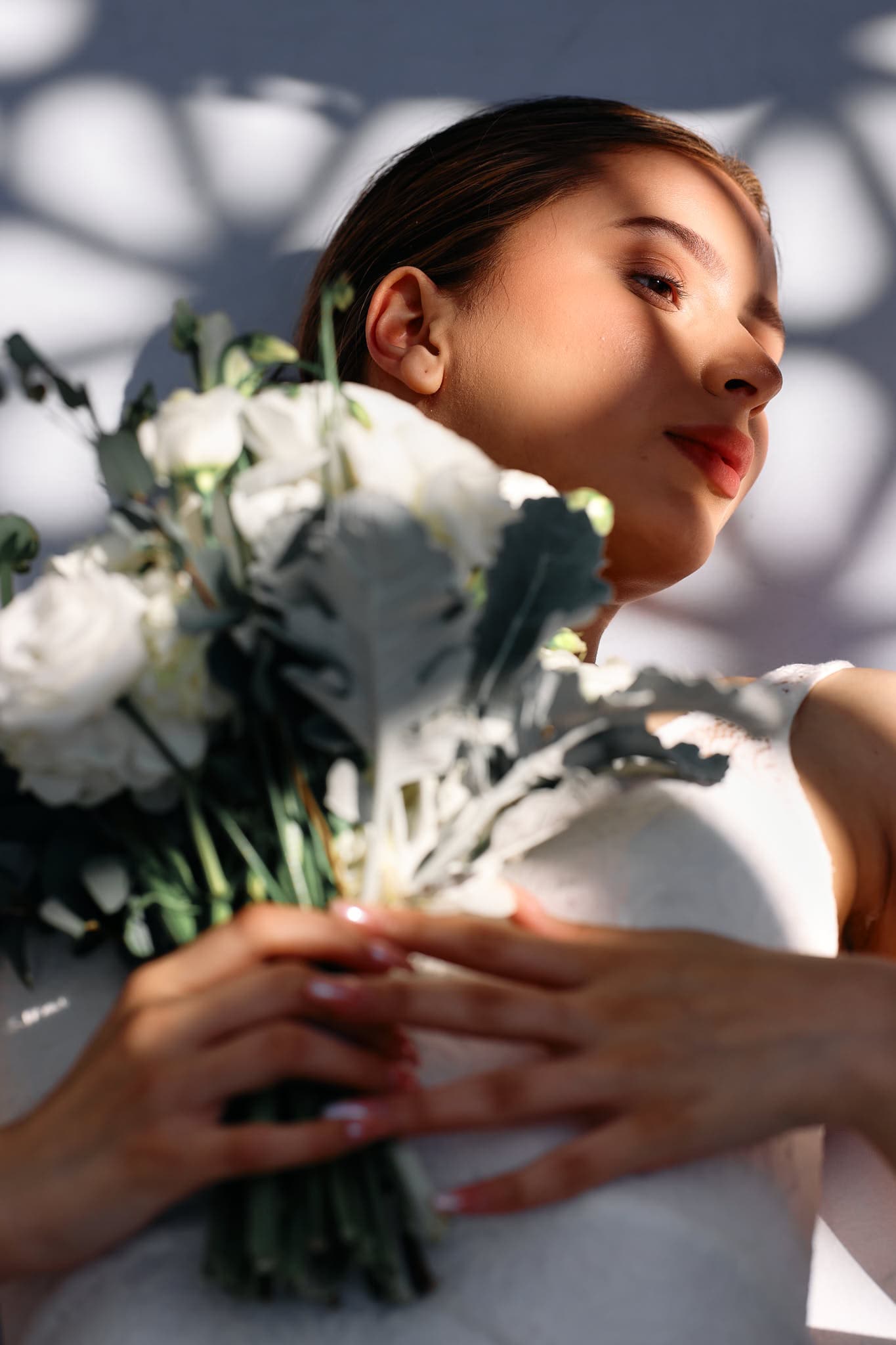 Delicate light patterns embellish this close-up of a bride holding a bouquet of fresh white roses and greenery. Soft lighting and delicate details highlight the elegance and romantic atmosphere, perfect for wedding and engagement photography.