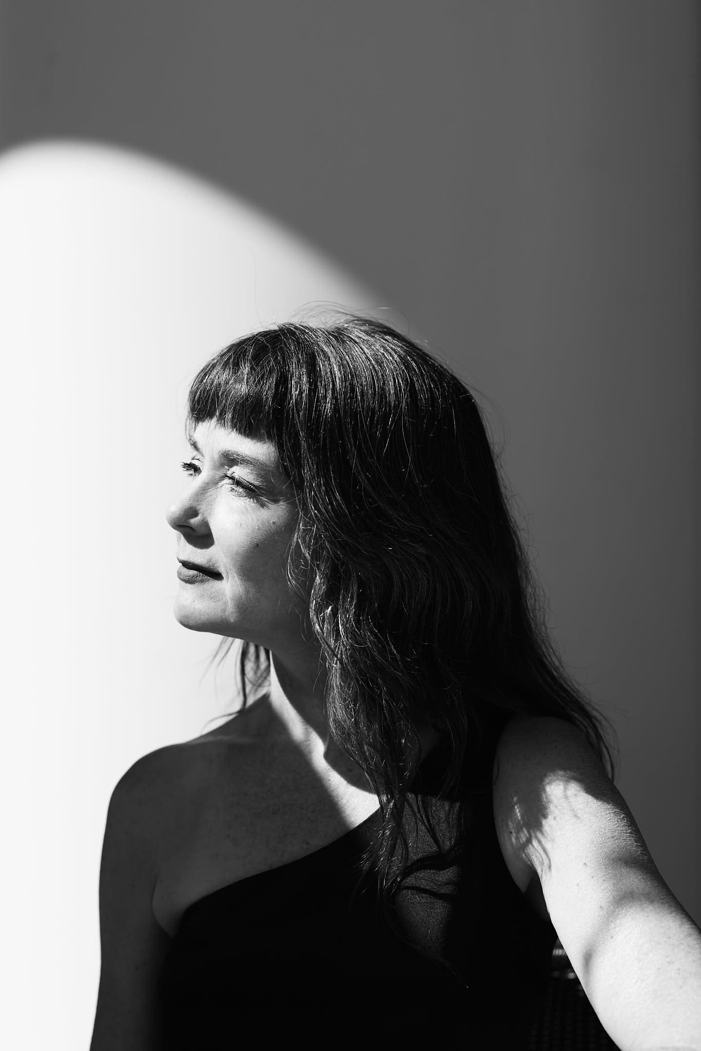Black-and-white portrait of a stylish individual with long, wavy hair, posing against a plain background. A dramatic one-shoulder top and bold shadows enhance the artistic, high-fashion aesthetic.