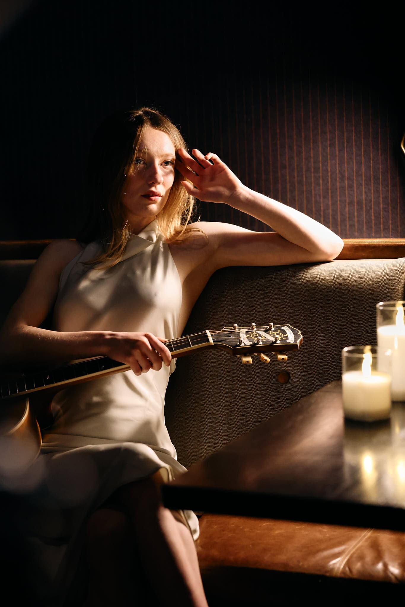Elegant portrait of a stylish woman seated on a cushioned bench, gently holding a guitar. Warm candlelight and dim surroundings enhance the intimate and artistic atmosphere, capturing a serene, soulful fashion editorial moment.