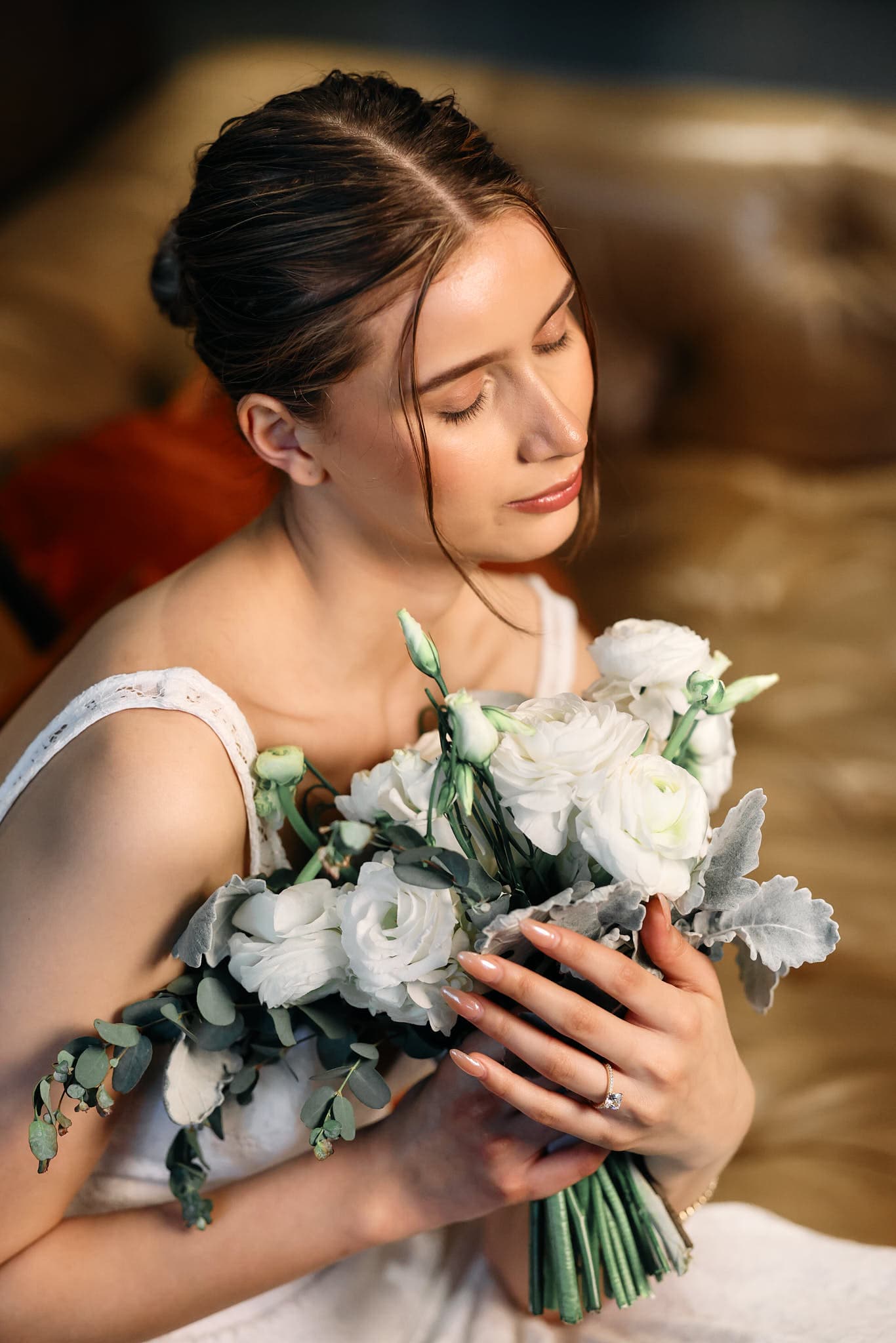Delicate close-up of a bride holding a bouquet of white roses and soft greenery. Her elegant sleeveless dress and sparkling engagement ring add to the timeless, romantic beauty of the moment, captured in soft, dreamy lighting.