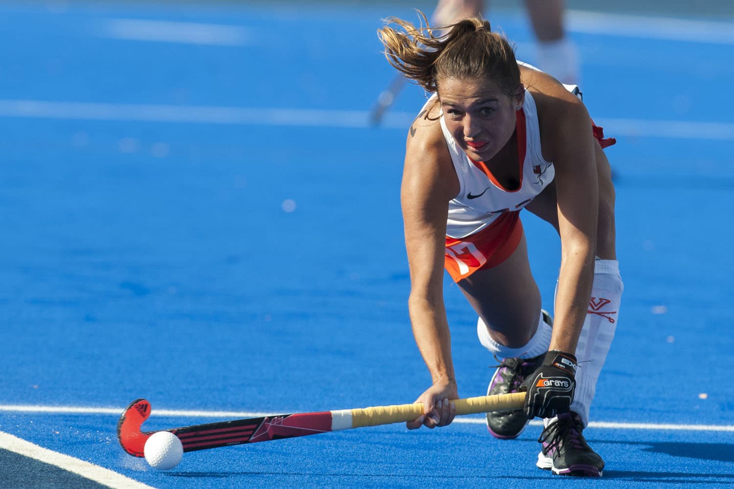 Virginia Cavaliers field hockey player Nadine de Koning (#17) lunges forward to make a pass during a match against Syracuse in Charlottesville, VA, on November 6, 2015. She grips her field hockey stick with both hands, aiming to send the ball across the blue turf. Dressed in a white and orange uniform, she wears protective gear, including gloves and shin guards, showcasing the intensity and athleticism of the game.