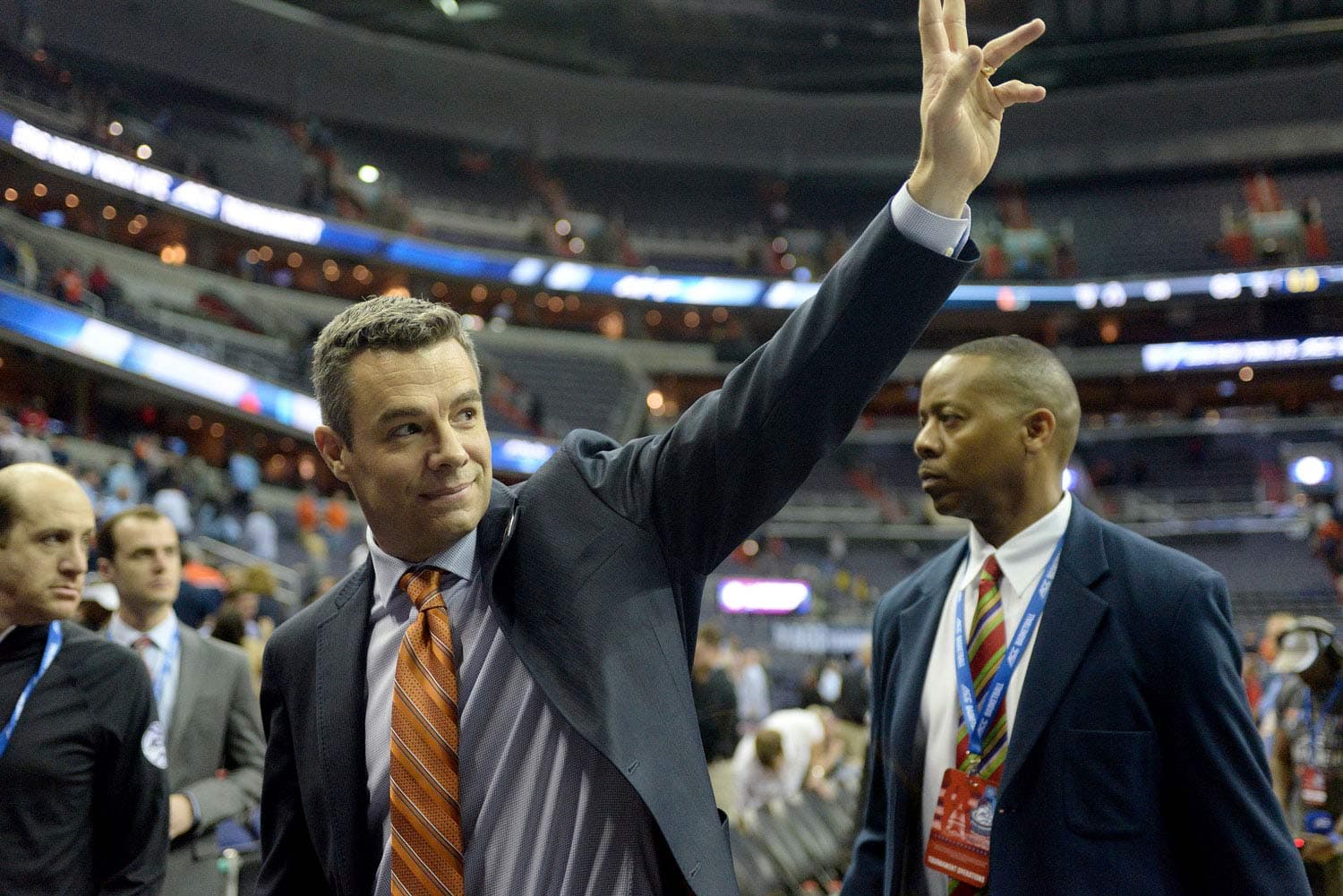 Virginia coach Tony Bennett waving to the crowd at the Verizon Center in Washington, D.C., after defeating Miami to advance to the ACC Basketball Tournament finals on March 11, 2016. Dressed in a dark suit, he raises his hand in appreciation as fans cheer in the background. The arena lights illuminate the scene, highlighting the celebratory atmosphere.
