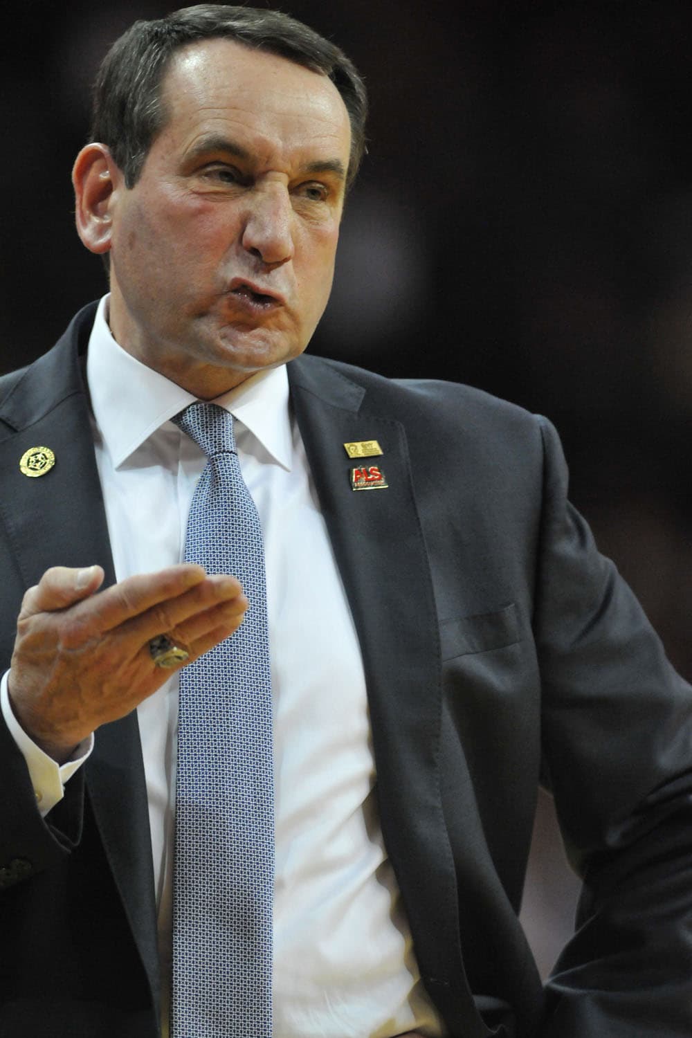 Coach K, dressed in a dark suit with a patterned tie, passionately arguing with a referee on the sideline during a basketball game. His right hand is raised in a gesture, emphasizing his point. Two pins are visible on his suit lapel, adding a distinctive touch to his attire. The background is dark and blurred, focusing attention on the intense exchange.
