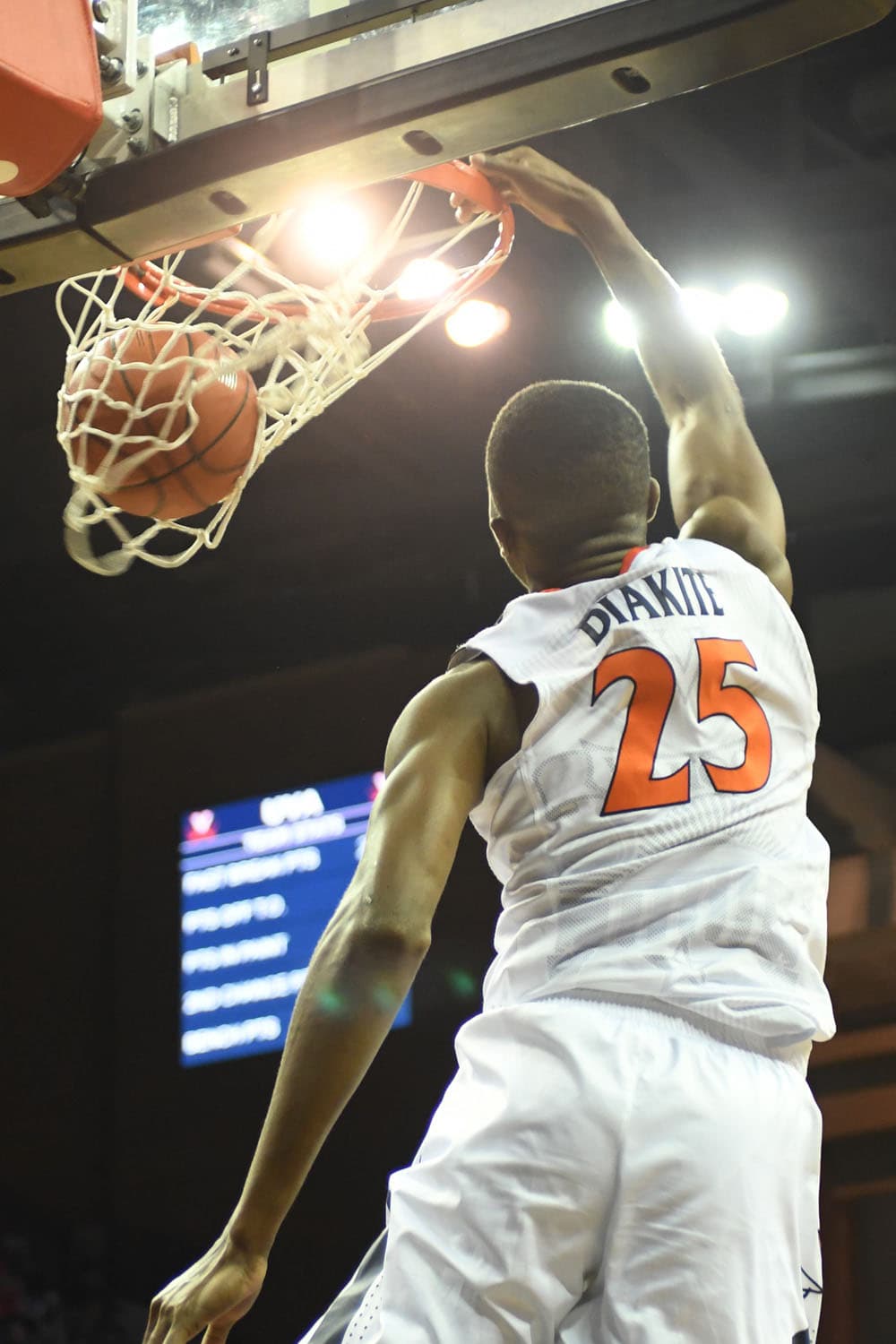 Mamadi Diakite of the Virginia Cavaliers, wearing jersey number 25, captured mid-air performing a slam dunk. The basketball is just above the hoop, emphasizing his athleticism and precision. The image is taken from a low angle, highlighting his height and dynamic movement. A blurred scoreboard is visible in the background, adding to the intensity of the moment.