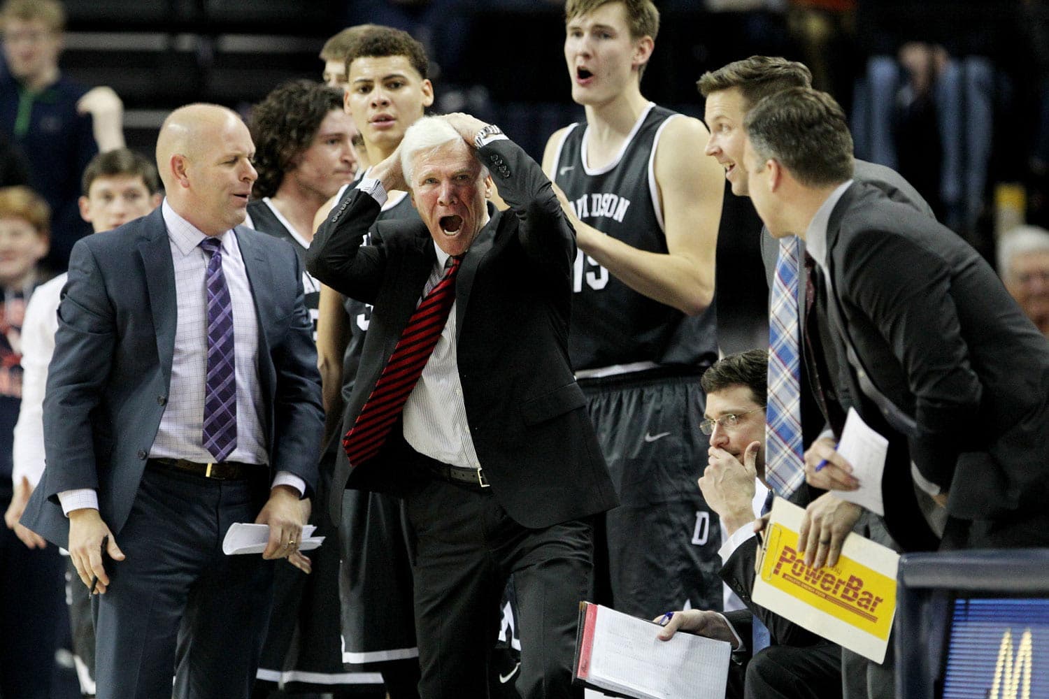 Davidson coach Bob McKillop, dressed in a suit with a red and black striped tie, visibly frustrated on the sidelines during the match against the Virginia Cavaliers at John Paul Jones Arena in Charlottesville, VA, on December 16, 2017. His hands are on his head, emphasizing the intensity of the moment. Surrounding him are Davidson players and staff, including one holding a clipboard and a PowerBar sign, adding to the scene’s competitive atmosphere.