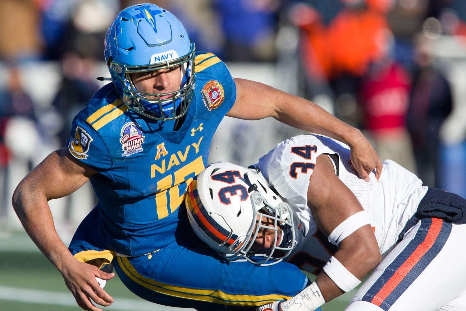 Navy quarterback Malcolm Perry (#10) being tackled by Kwontie Moore (#34) of the Virginia Cavaliers during the Military Bowl at Navy-Marine Corps Memorial Stadium in Annapolis, MD, on December 28, 2017. Perry, wearing a blue Navy jersey with Military Bowl patches, is brought down in an intense moment of action. The stadium lights illuminate the field, highlighting the competitive atmosphere of the game.