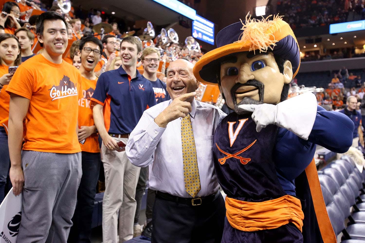 Dick Vitale, legendary sports commentator, standing courtside at a Virginia Cavaliers basketball game. He is seen engaging with fans, with the Cavaliers mascot nearby, dressed in a navy blue outfit with an orange sash and a large hat. The crowd in the background is animated, with some holding musical instruments, indicating the presence of a band.