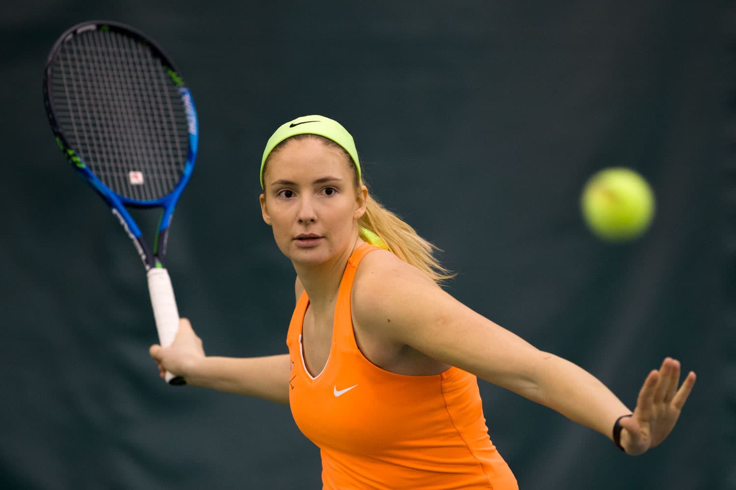Teodora Radosavljevic of the Virginia Cavaliers preparing to unleash a powerful forehand at the Boar's Head Sports Club in Charlottesville, VA, on January 18, 2018. She is dressed in a navy and orange uniform, gripping her racket with precision as she focuses on the incoming ball. The indoor court setting highlights the competitive atmosphere of the match.