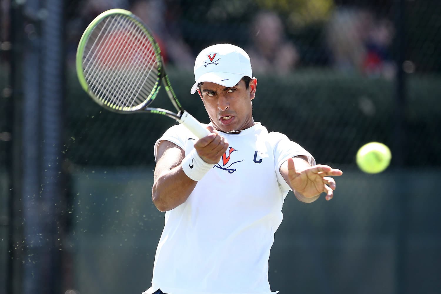 Aswin Lizen of the Virginia Cavaliers executing a powerful volley against the NC State Wolfpack at Snyder Tennis Center in Charlottesville, VA, on April 13, 2018. Dressed in a white and navy uniform, he focuses intently on the ball, showcasing precision and athleticism. The court surface and surrounding fencing frame the action, emphasizing the competitive atmosphere of the match.