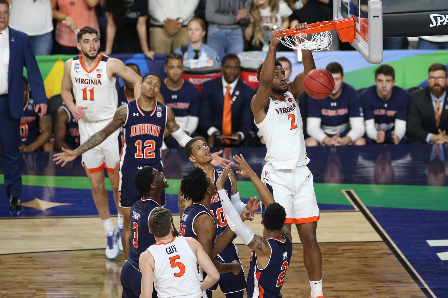 Braxton Key of the Virginia Cavaliers, wearing jersey number 2, captured mid-air performing a slam dunk during the 2019 Final Four game. The basketball is in his grasp as he hangs onto the rim, showcasing his athleticism. Auburn Tigers players, dressed in navy blue jerseys with orange accents, are positioned around him, attempting to defend. The crowd and other players are visible in the background, adding to the intensity of the moment.