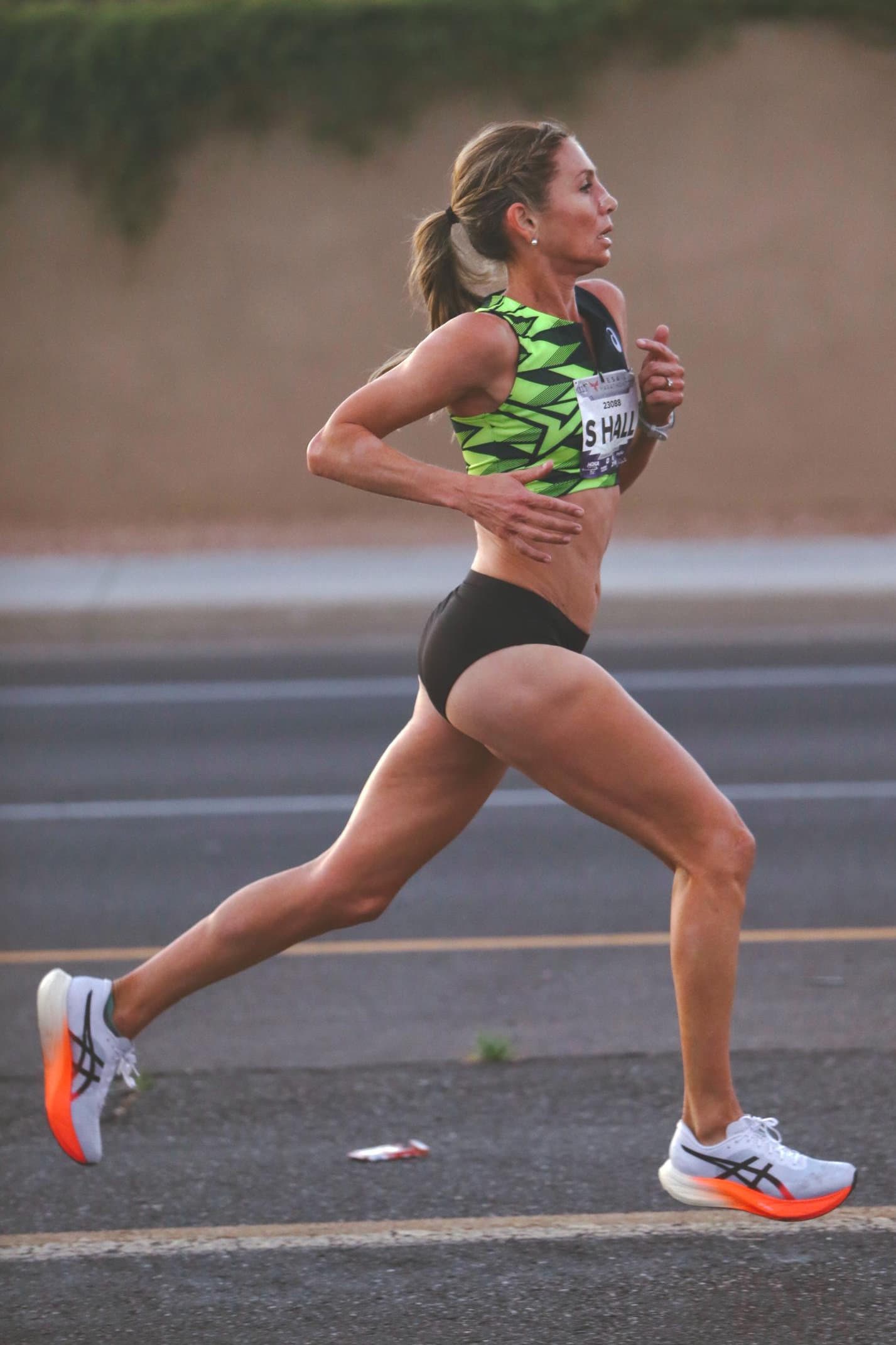 Former steeplechase champion Sara Hall of the United States strides six miles into the Mesa Half-Marathon on February 8, 2025, in Mesa, Arizona. Wearing a black and forest green racing crop, black speed buns, and white with orange accented ASICS super shoes, her powerful stride and focused expression reflect endurance and elite performance in this dynamic marathon moment.