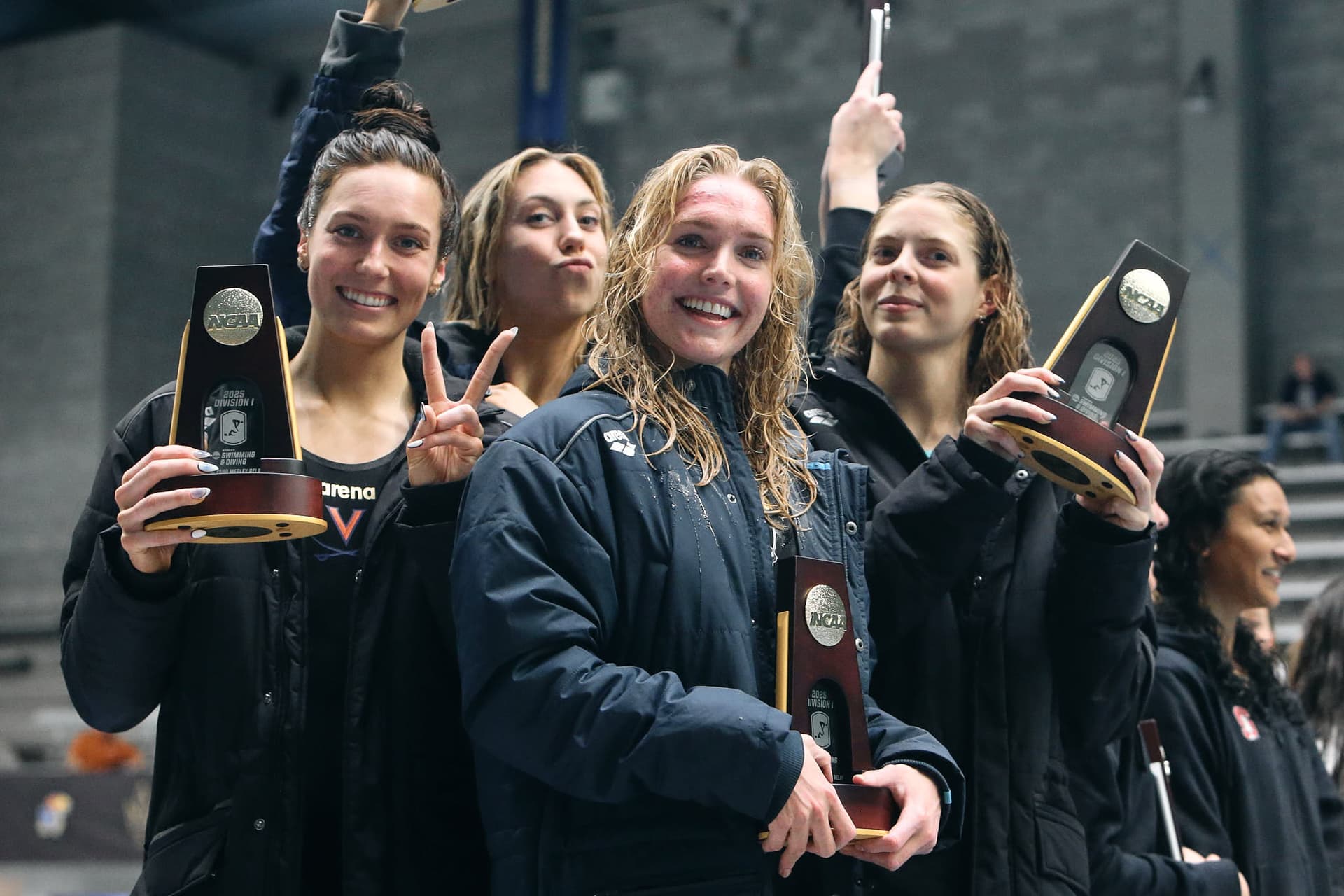 Virginia Cavaliers women's 200-yard Medley Relay National Champions celebrating their victory at the NCAA Championships. The athletes, dressed in dark jackets, proudly hold their NCAA trophies, which display the text 'NCAA' and 'Division I.' One team member flashes a peace sign, adding a personal touch to the moment. The indoor setting suggests a formal awards ceremony, capturing the excitement and achievement of the team.
