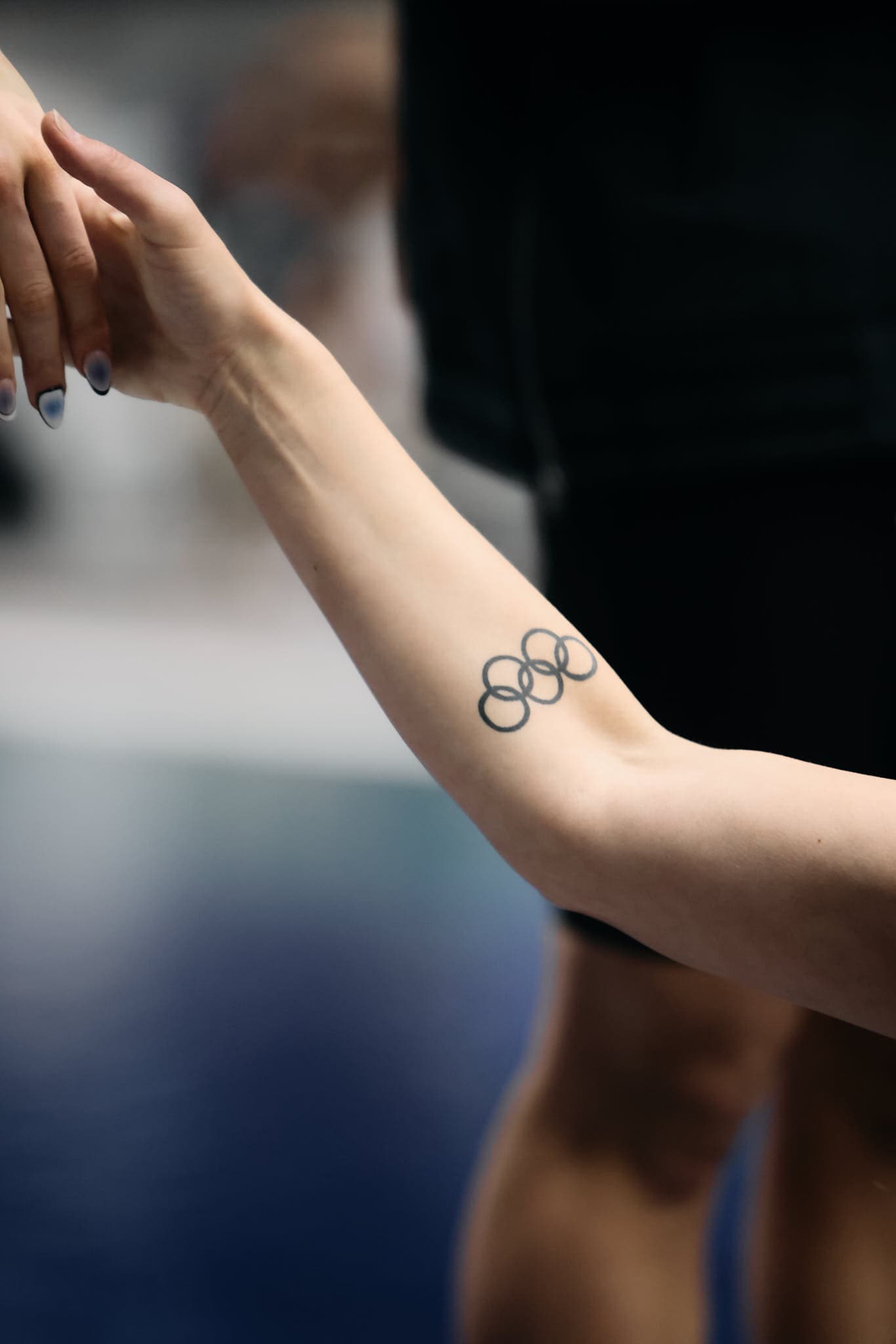 An athlete receiving a handshake from a former Olympian during an awards ceremony at the Weyerhaeuser King County Aquatics Center in Federal Way, Washington, on March 20, 2025. The Olympian's arm features a tattoo of the Olympic rings, symbolizing their past achievements. The handshake captures a moment of recognition and celebration in a competitive sports setting.