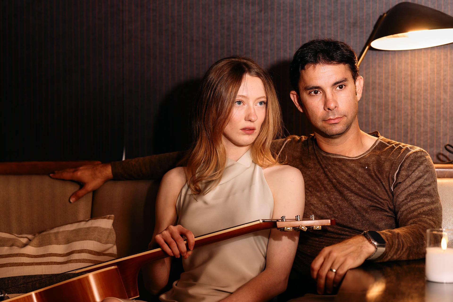 Stylish couple posing intimately on a couch for a fashion editorial shoot. The man holds a guitar, adding an artistic touch, while the warm lighting and cozy setting emphasize a modern, relaxed elegance.