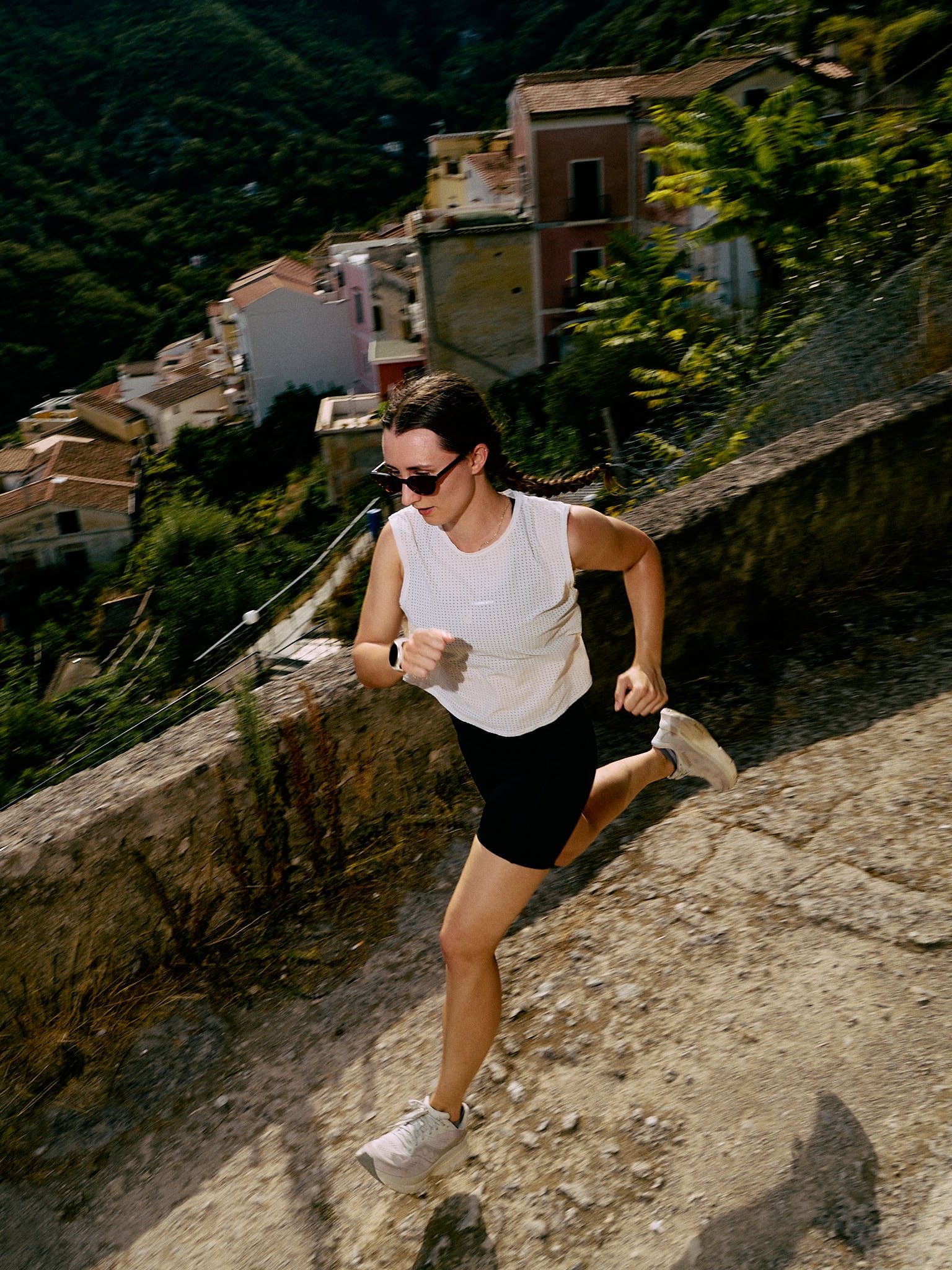A woman runs uphill along a stone path past hillside houses and greenery in Vietri sul Mare, Italy.